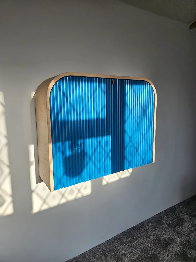 Wall-mounted fold-down desk, closed, with a curved timber case and a blue fluted front